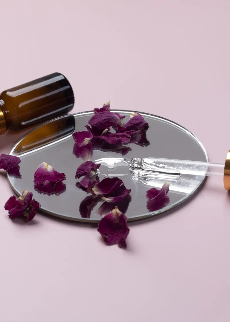 A glass pipette and amber serum bottle on a mirror, surrounded by dried rose petals